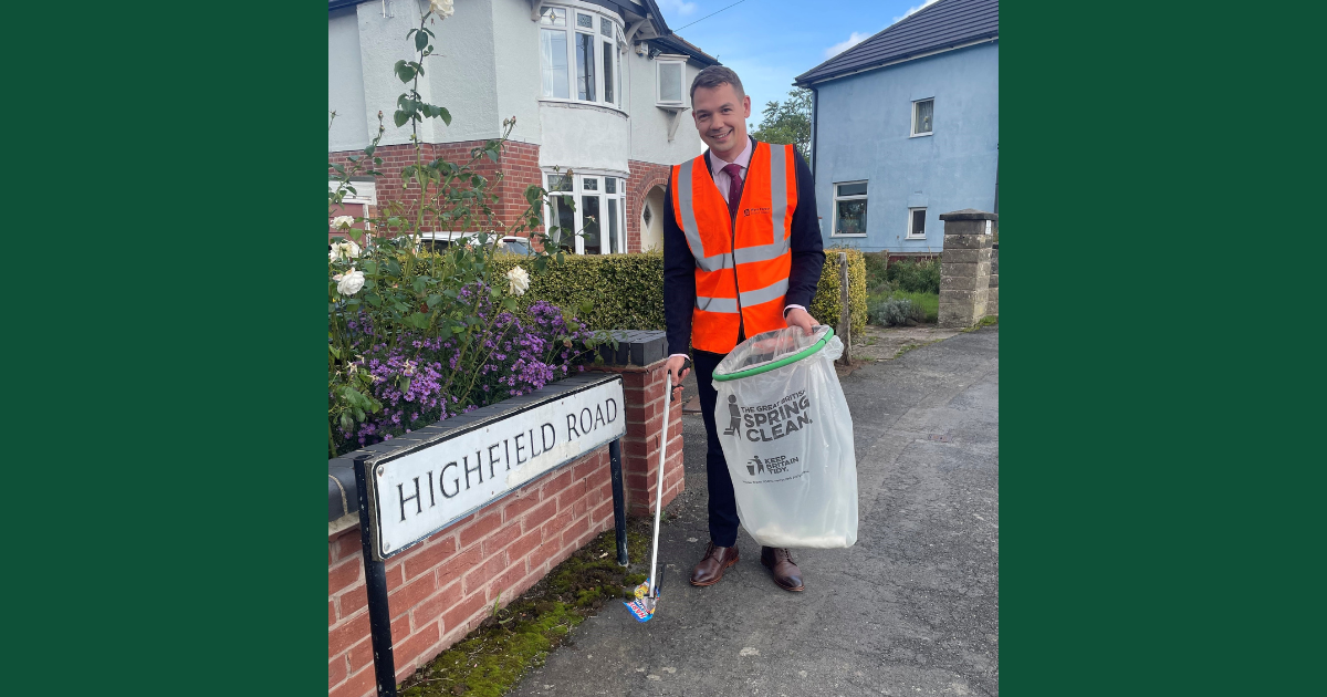 Councillor Ben Brookes in orange hi-viz jacket litter picking next to street name with picker and bag