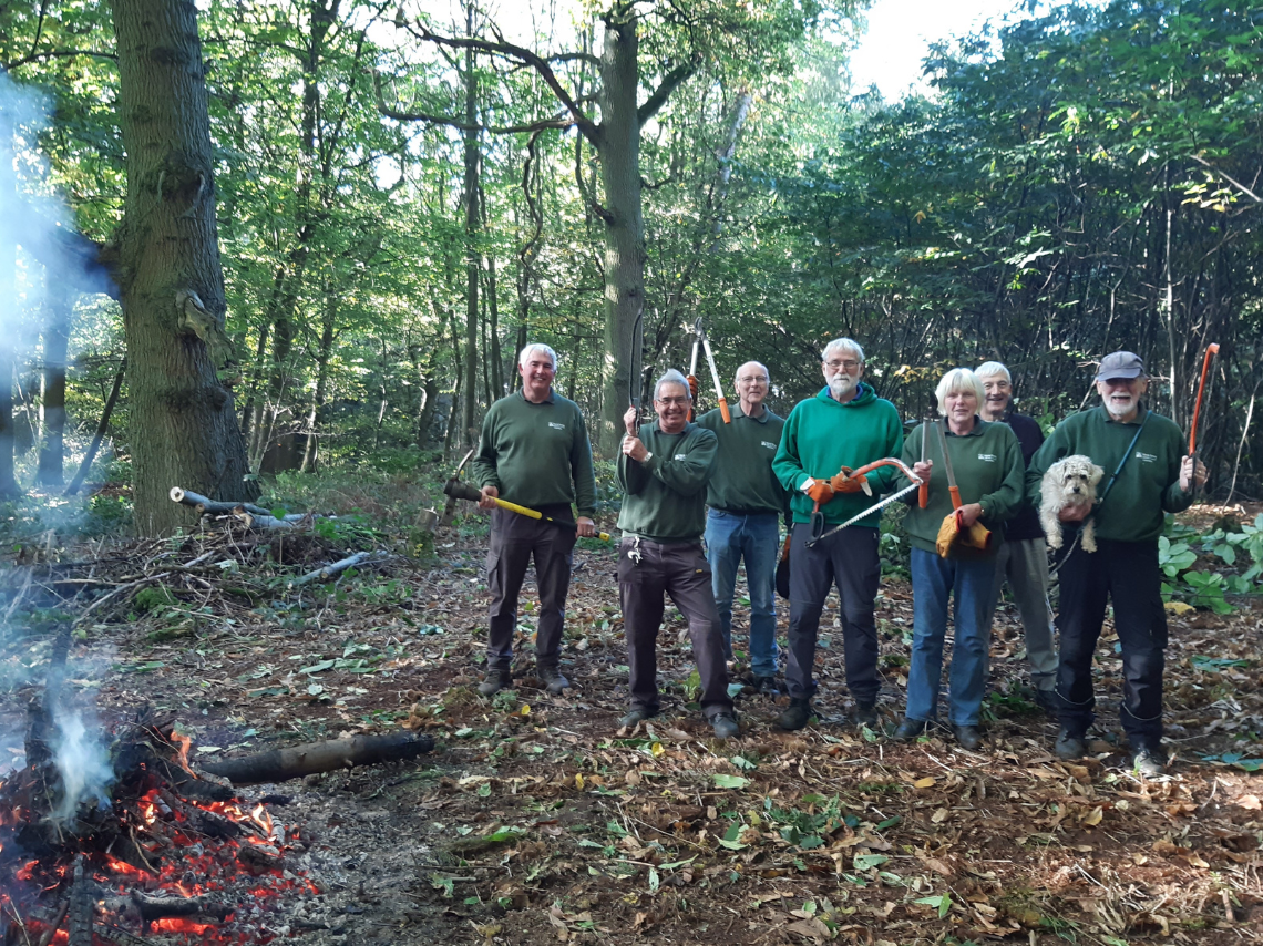 6 men and one woman wearing mostly green in a wooded area carrying tree cutting tools.  Open fire to left of picture and man holding a white fluffy dog on the right.
