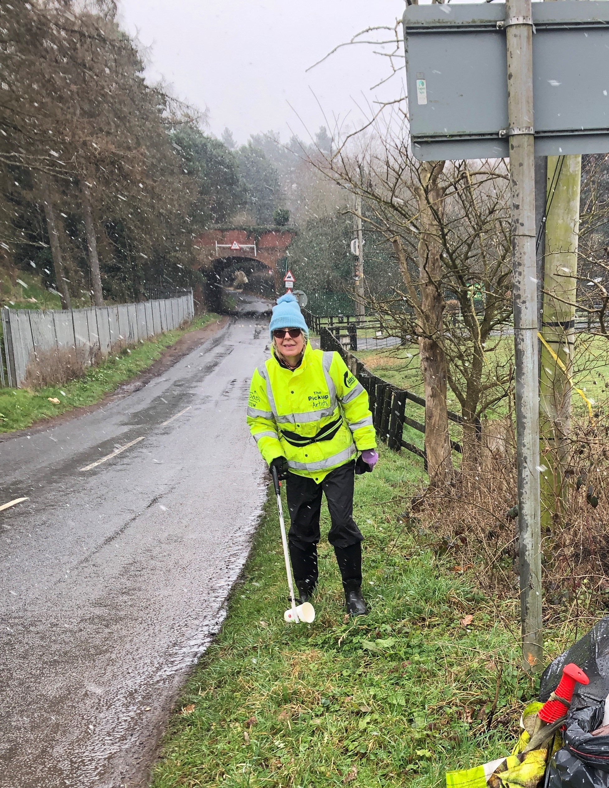 A woman in high visibility jacket litter picking on the roadside with snow falling