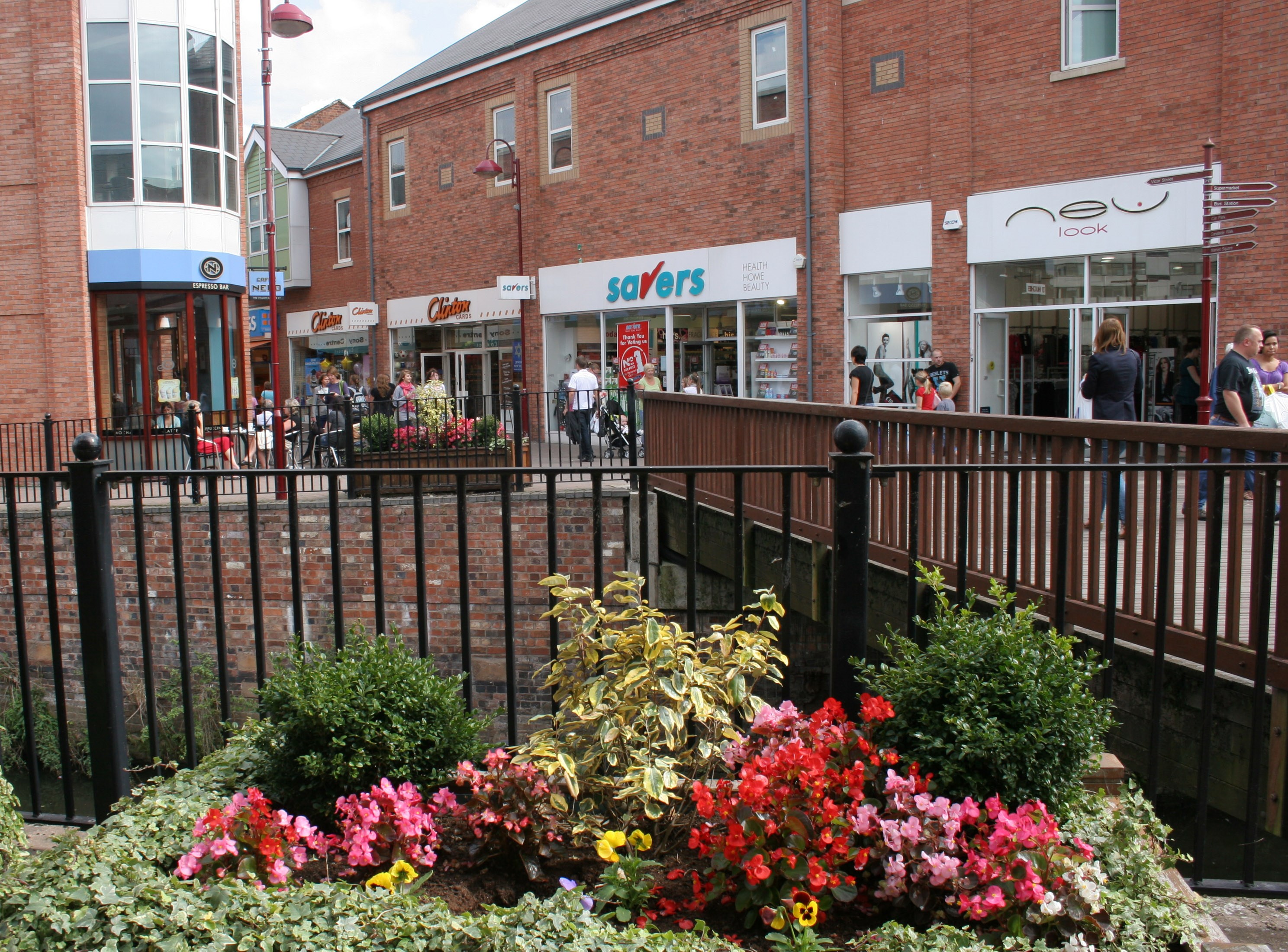 in the foreground there are several variations of summer bedding plants of various colours such as, reds, yellows and pinks. In the background is a local town high street with pedestrians shopping in th elocal stores. 