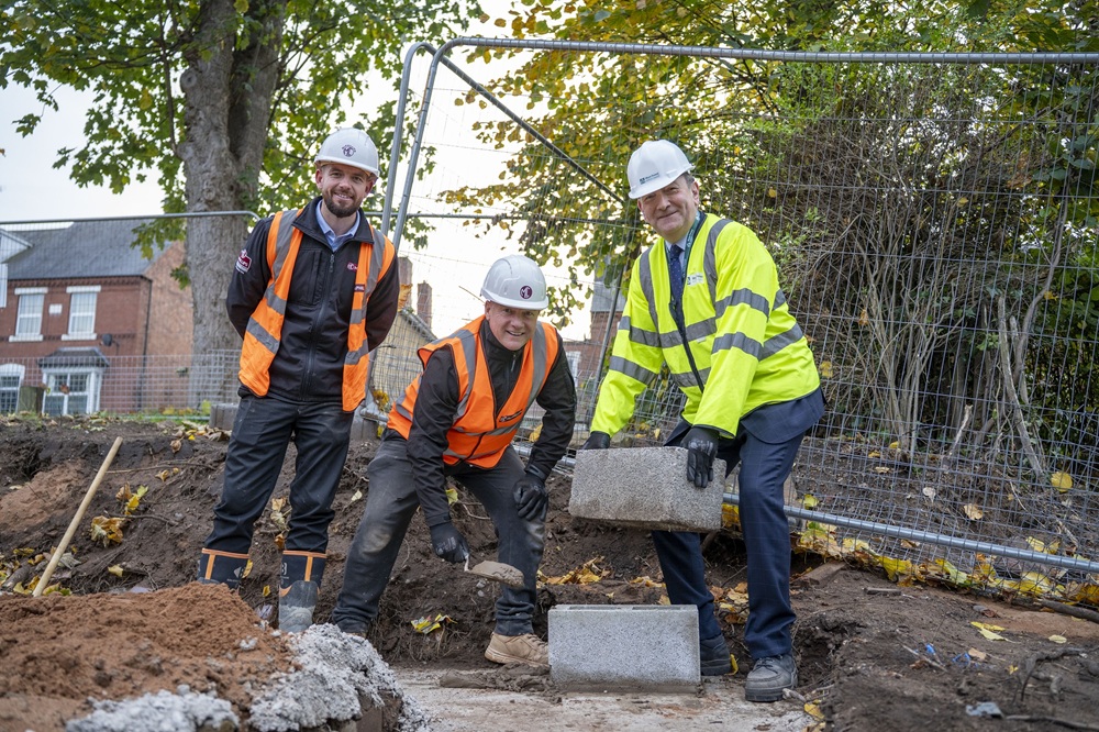 Three people wearing high visibility jackets, hard hats and gloves place a brick on a building site