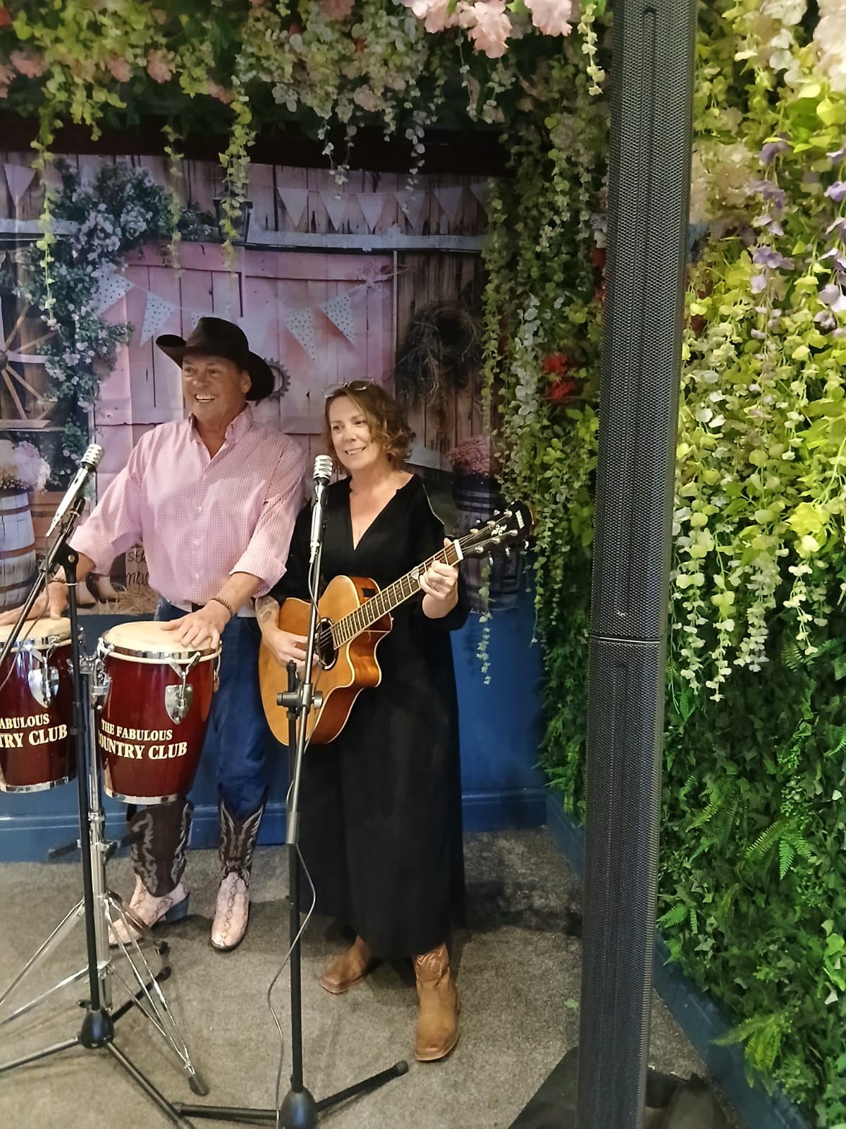 Photo of two musicians in an outdoor setting, one male in a pink shirt, wearing a cowboy hat playing some drums by hand. the other musician is female in a black dress playing a guitar