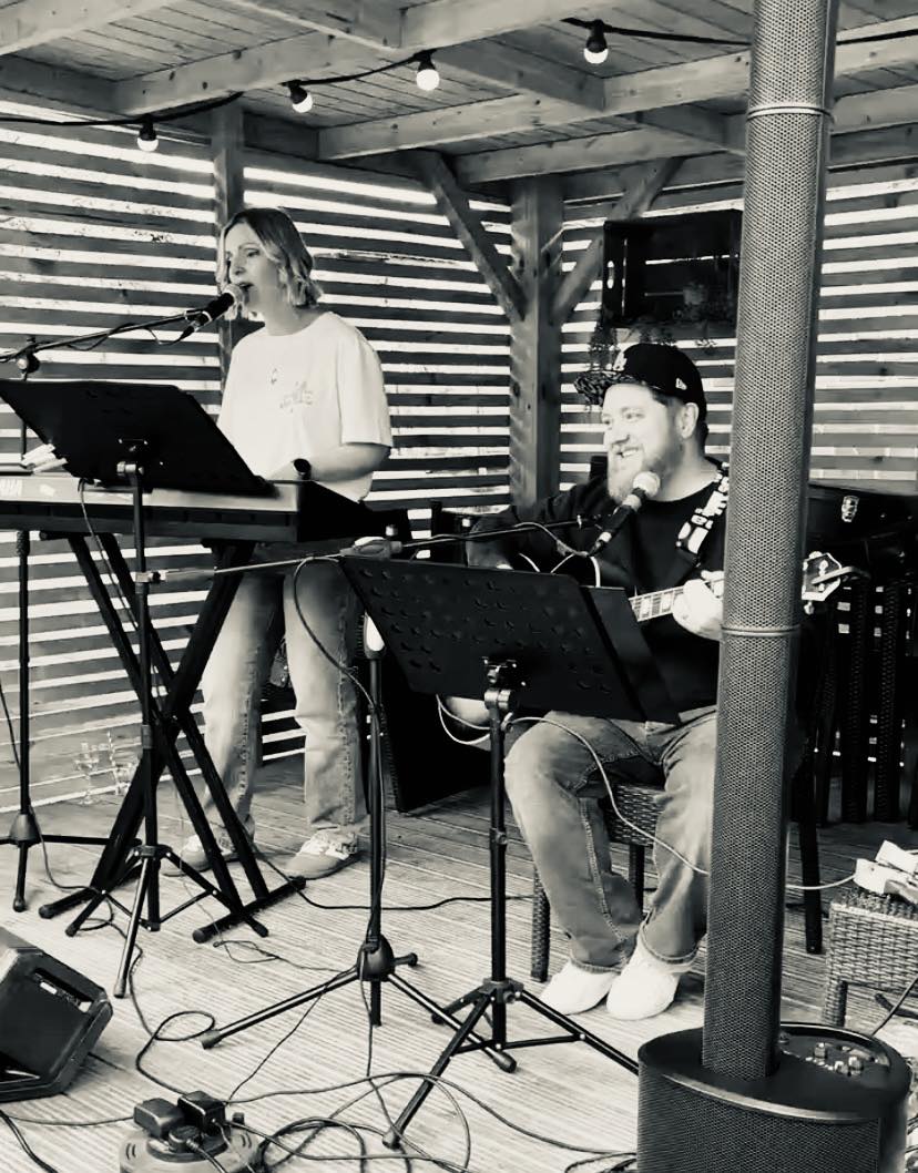 Black and white photo of the two members of the group Friday Club. One is playing the keyboard and singing. The second is playing the guitar and singing.