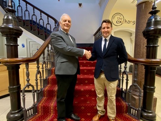 Two males wearing business attire stood at the bottom of a grand staircase with a rich red carpet and wrought iron balustrades shaking hands