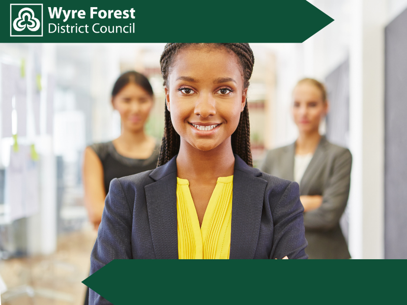 A young girl in business wear smiling, there are two blurry people in business wear stood further back behind her.