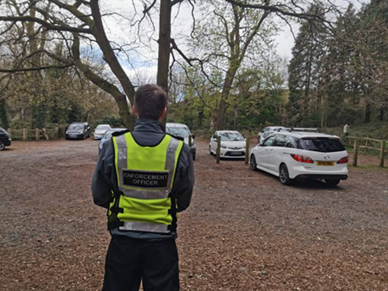 Photo taken from the back of a person wearing a high visibility jacket that says Civil Enforcement, they are looking over a car park with parked cars in a rural area
