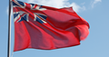 The Red Ensign Flag ( red background with the Union Jack in the top left corner) raised on a flagstaff with a clear blue sky in the background