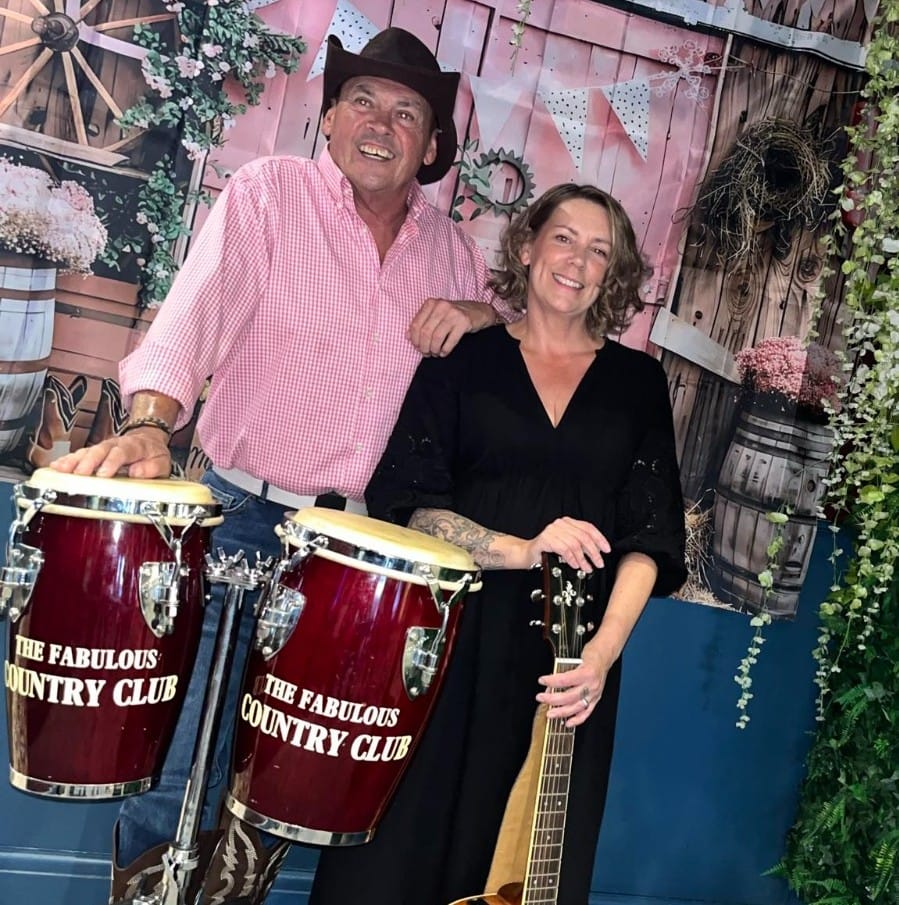  Photo of two musicians, one male in a pick shirt wearing a cowboy hat and one female wearing a black top. Both are stood in front of two dark red drums
