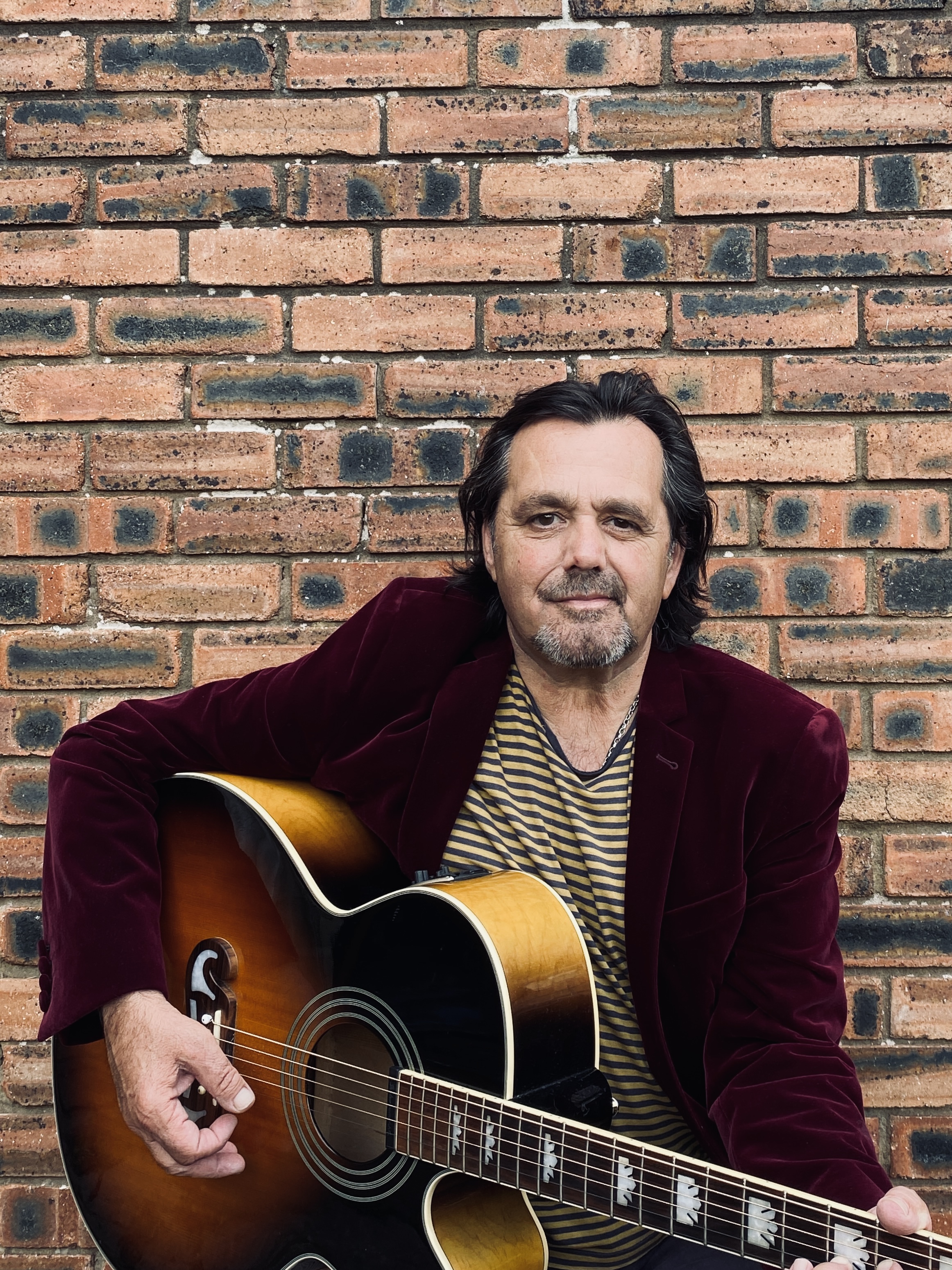   Photo of a male performer standing against a brick wall holding a guitar