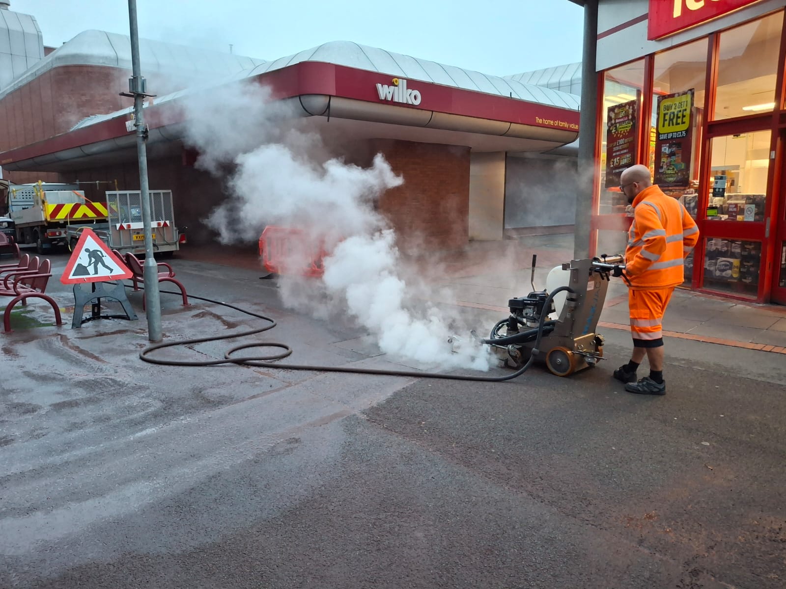 A worker dressed in orange high vis gear using a chewing gum remover on the pavement outside shops in a town centre. 