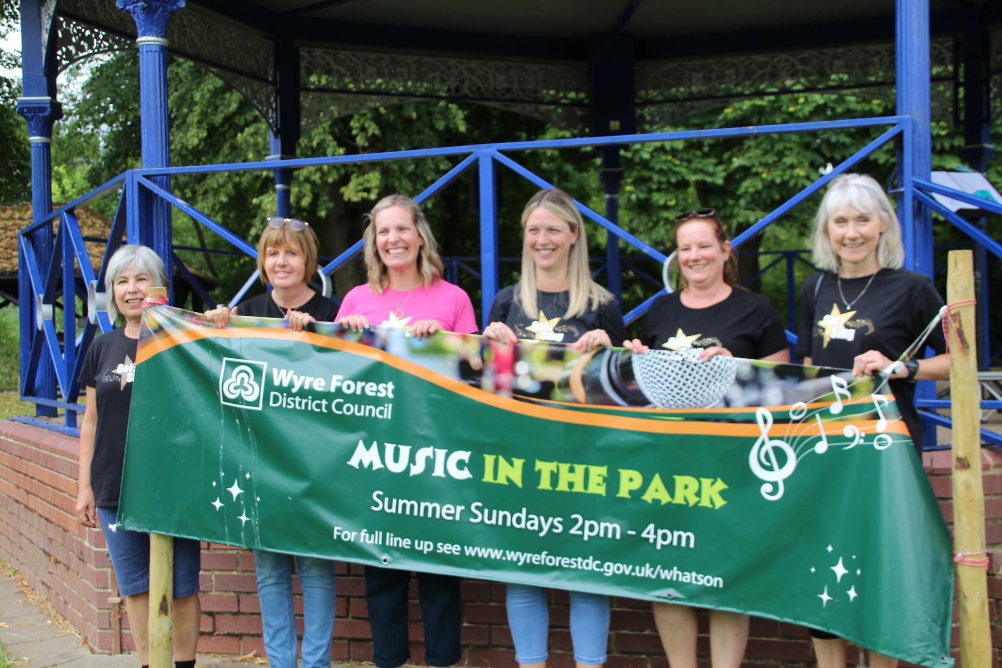Six women members of the choir standing in front of the bandstand at Riverside Meadows. They are holding the council banner which has Music in the Park words.  