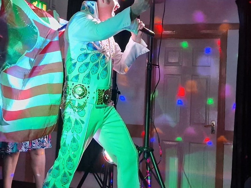 Photo of an Elvis Presley tribute singer - holding and singing into a microphone in front of an American flag