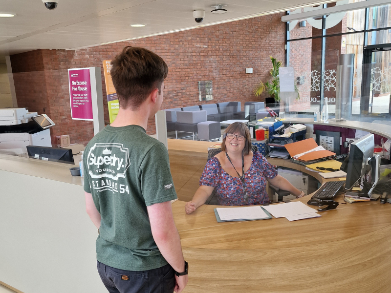a male stood at a reception desk with a female receptionist smiling back