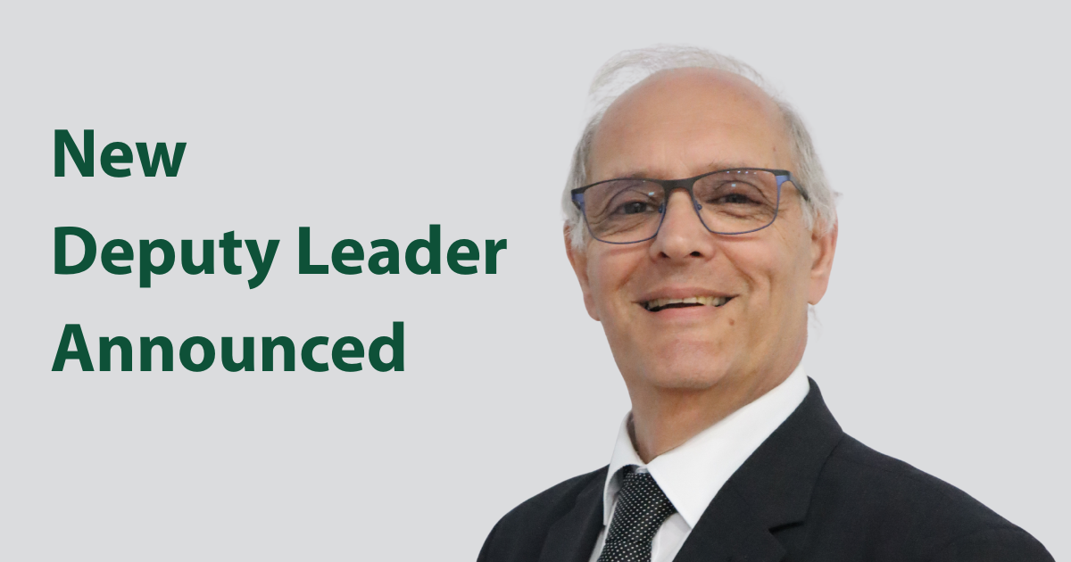 Portrait photograph of balding man with grey hair and glasses. He is wearing a dark suit and tie