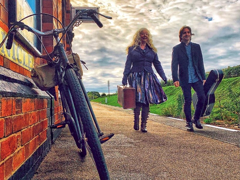 Image of a woman and a man walking together outside. The woman is carrying a small suitcase. The man is carrying a guitar case. In the front of the image is a bicycle leaning up against a brick building