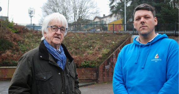 Two men, one with white hair and glass, the other wearing a bright blue hoodie stood in an underpass