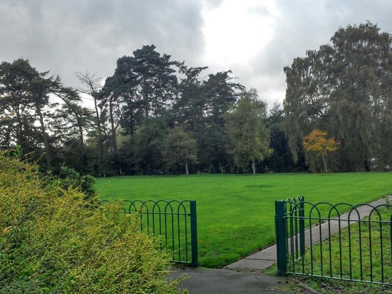 A beautiful lawned area bowling green surrounded by black metal railings, with mature trees that be seen in the distance that border the grassed area.