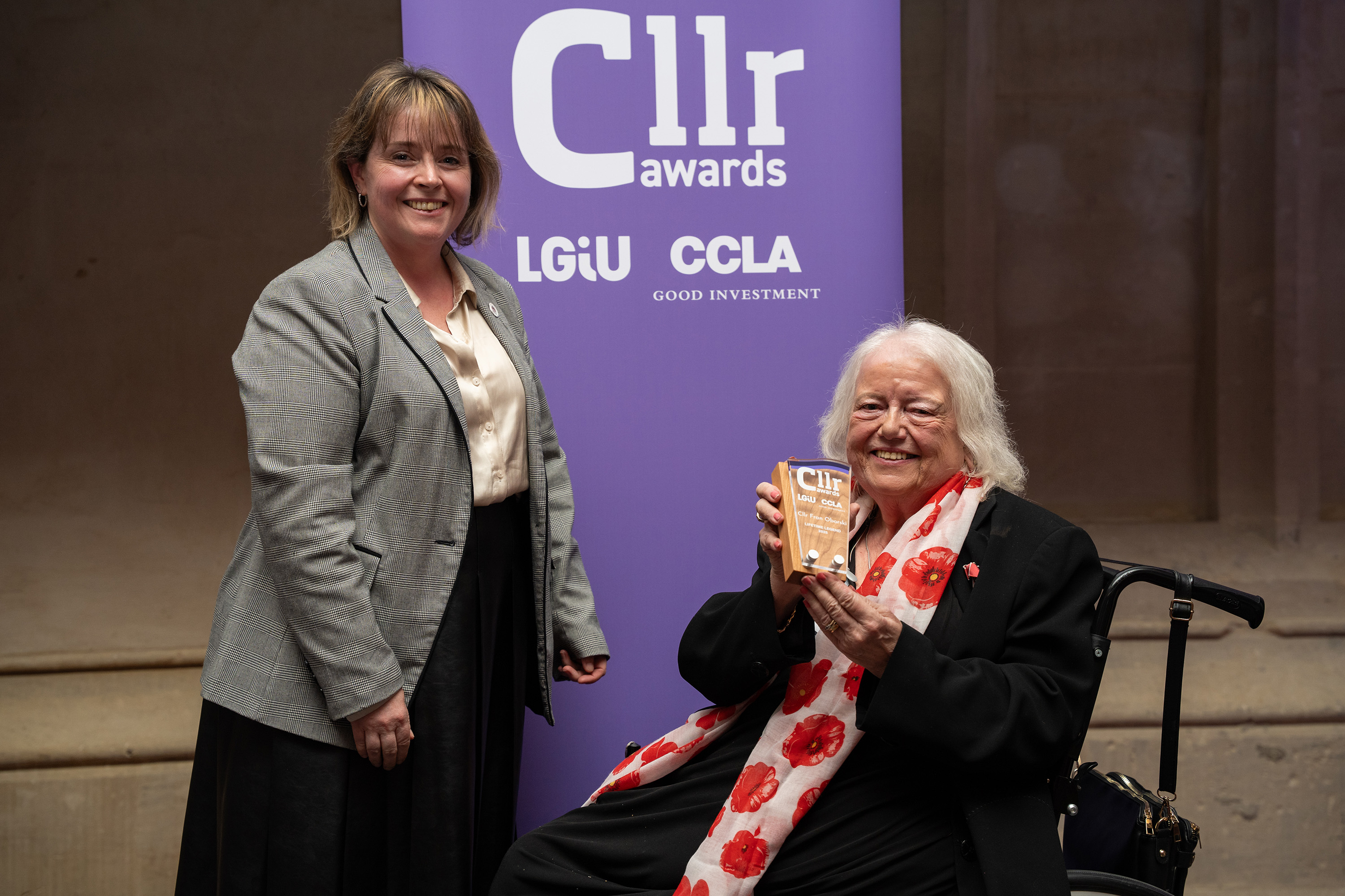 Two women side by side in front of a purple banner which says Cllr Awards, one is standing and the other is in a wheelchair holding an award trophy