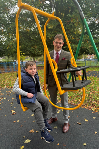 Man wearing  suit and glasses stood by boy wearing a jeans and navy puffer gillet. Boy is sat on one side of a two seated swing.