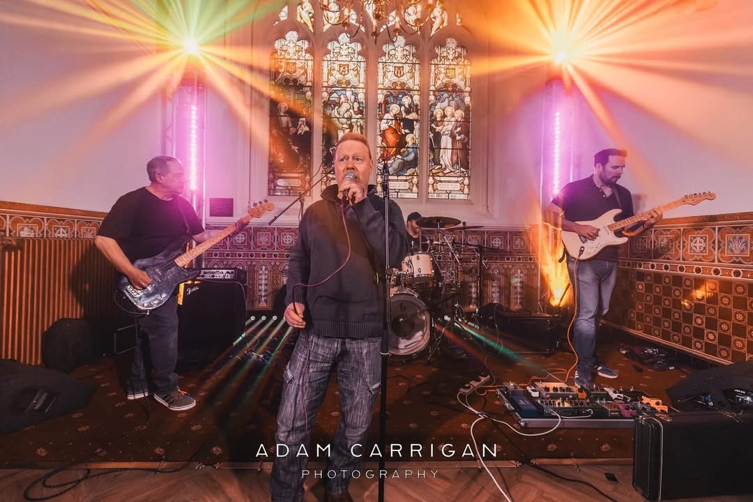   Photo of the 3 male members of the Cannon Beach Band, two are playing guitar and one in the centre of photo is singing. the are in church like environment with a stained glass window behind