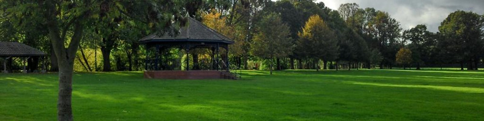 Open green space with a large established tree in the foreground and a bandstand in the background