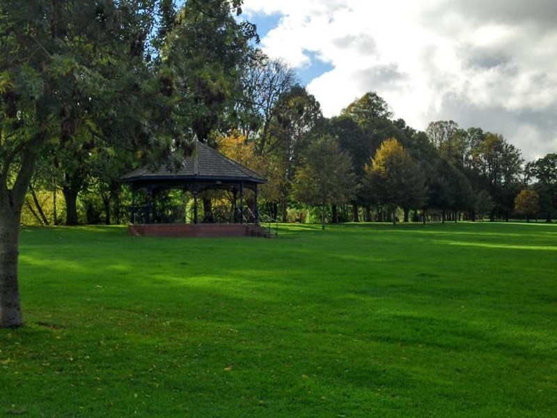 A rassed area with a bandstand located against a backdrop of mature trees which border the park.