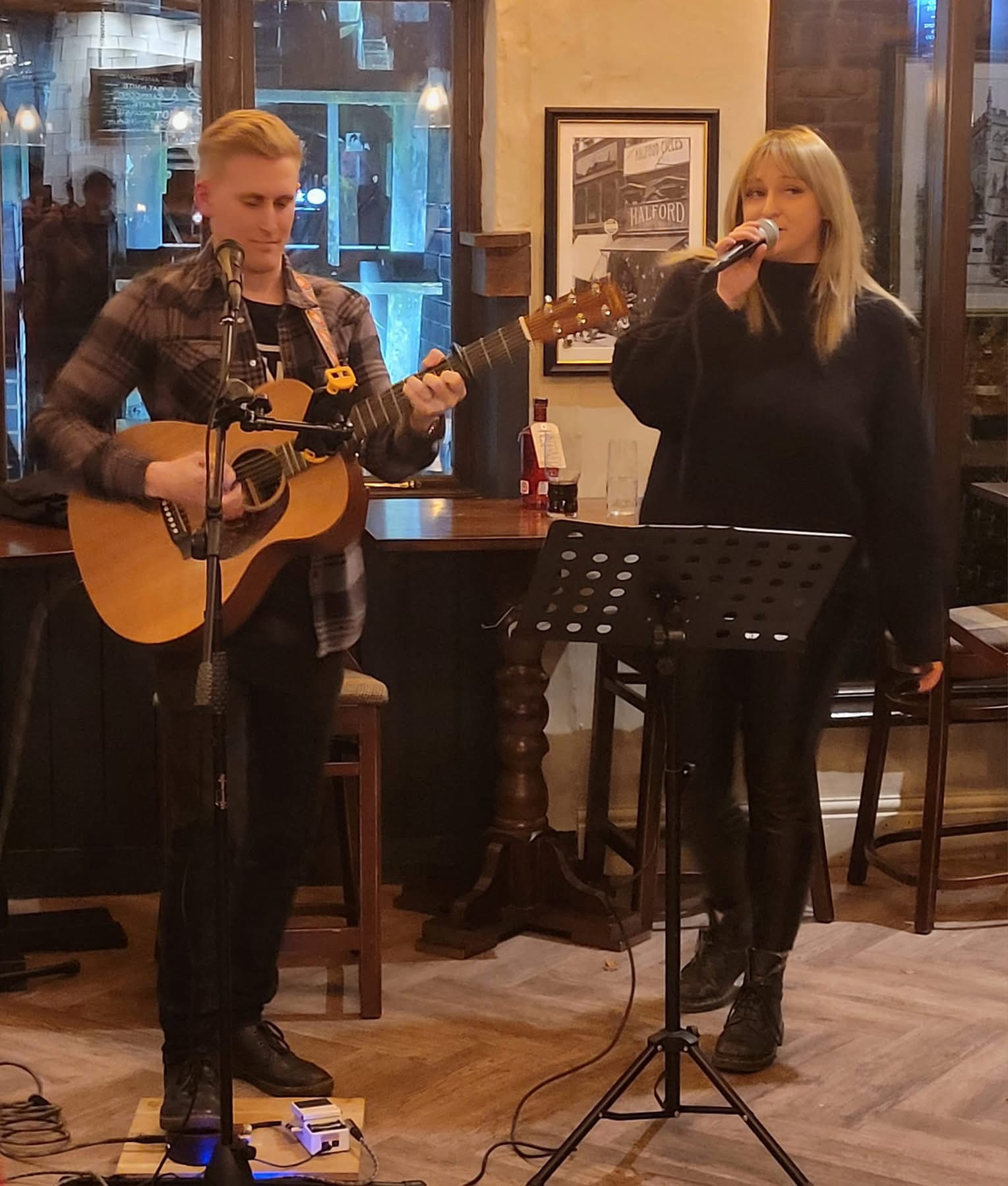   Photo of the 2  performers of the acoustic duo Milky and Liv. One is playing the guitar and one is holding a microphone and singing. they are in pub setting.