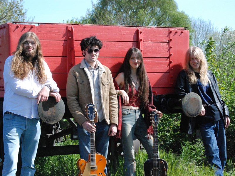 Image of the band Crimson Band, 3 male musicans and one female performer stood outside leaning against a red trailer.