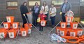 5 adults and one child standing in front of a wooden scout hut smiling and holding litter pickers, the child is holding an orange bucket, there are orange buckets stacked in a triangle formation either side of the group 