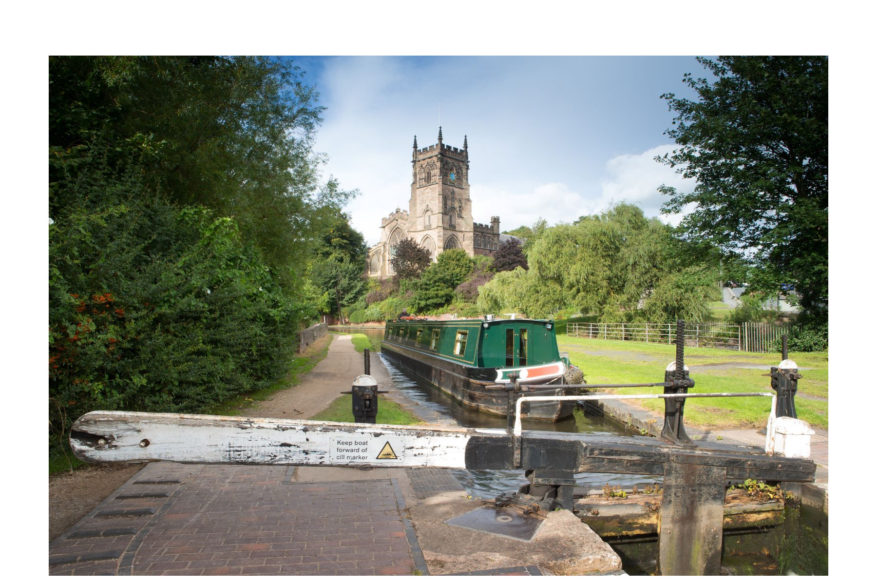 A distant view of St Mary's Church across the local canal. The canal mirrors a reflection of the church across the top of the water. Trees line the canal towpath on the left side.