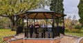 Image of a metal bandstand with a brick base and access ramp in a park, with four people standing inside.