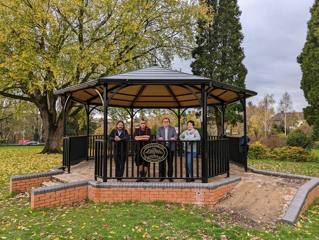 Image of a metal bandstand with a brick base and access ramp in a park, with four people standing inside.