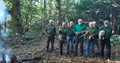 6 men and one woman wearing mostly green in a wooded area carrying tree cutting tools.  Open fire to left of picture and man holding a white fluffy dog on the right.