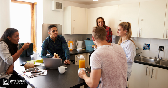 Group of young people sat around a kitchen breakfast bar. One has a lap top.