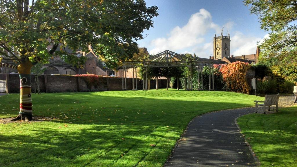 Open air theatre in public gardens surrounded by trres and ivy growing up a wall with the local church in the distant background