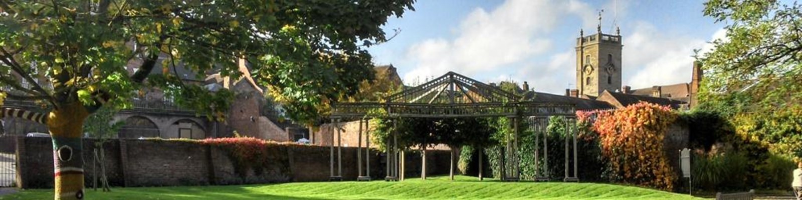 Open air theatre in public gardens surrounded by trres and ivy growing up a wall with the local church in the distant background