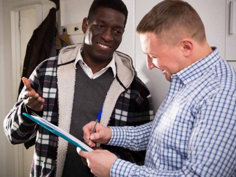 Two smiling people stood in house, one person is writing on clipboard