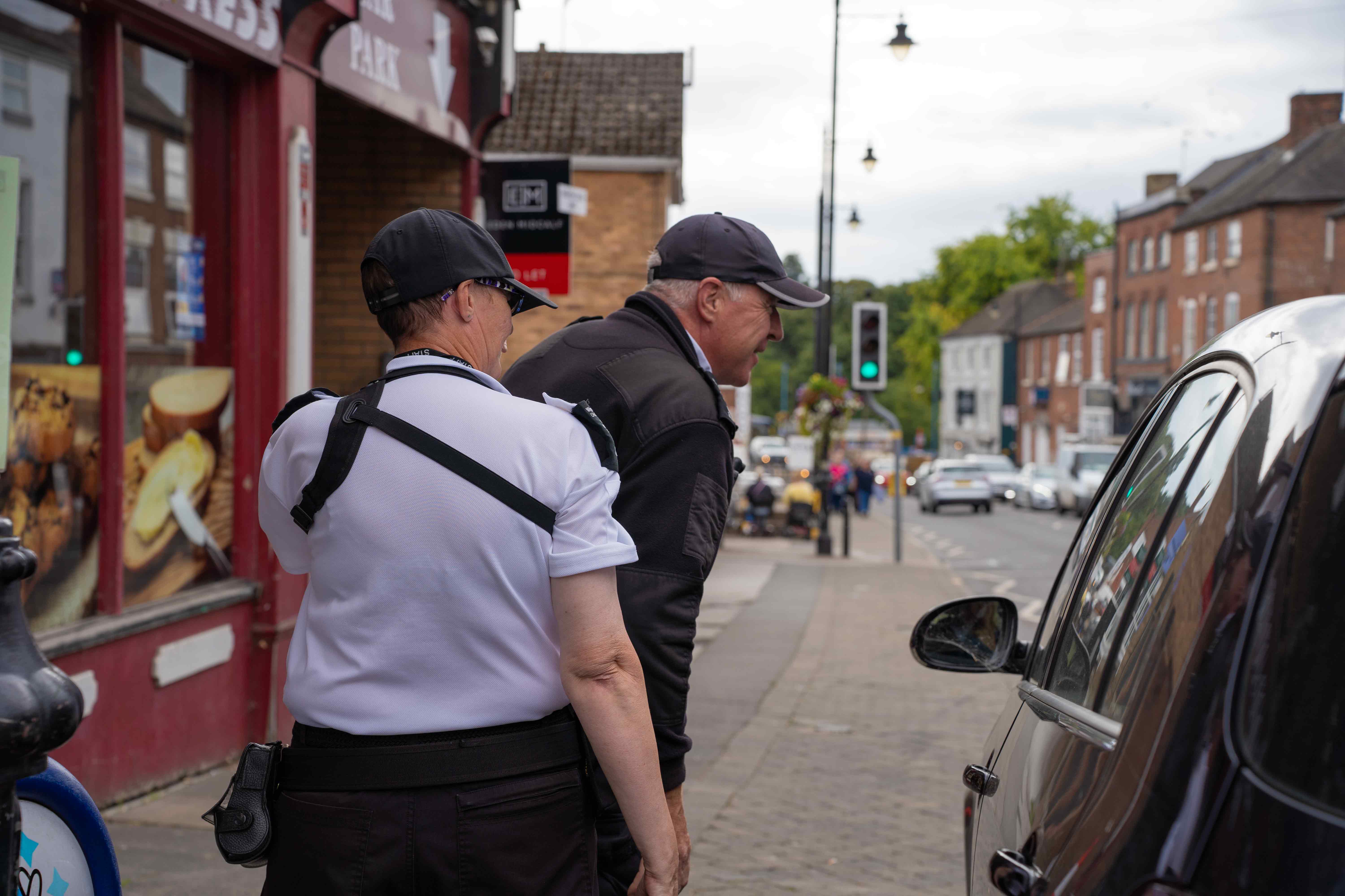 Two people in enforcement officer uniforms, leaning towards a car to talk to someone inside. 
