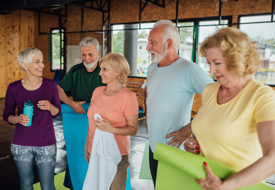 A group of five older people standing together smiling and holding water bottles, towels and yoga mats