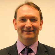 headshot of man with dark side-parted hair and a mole above right eyebrow