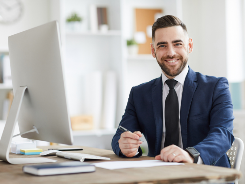A man in a dark blue suit smiling, he is sat at a desk in front of a computer