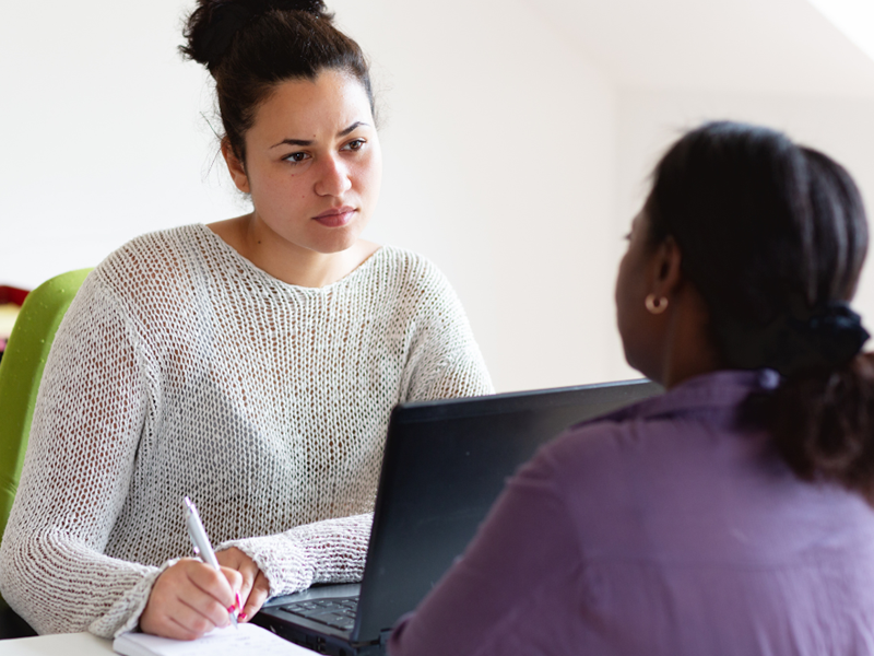 Two people sat across a desk from each other, one person is listening and taking notes in a notepad.