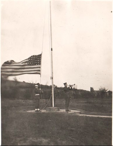 a soldier lowers US flag while another plays bugle at flag pole in field. Old sepia photo